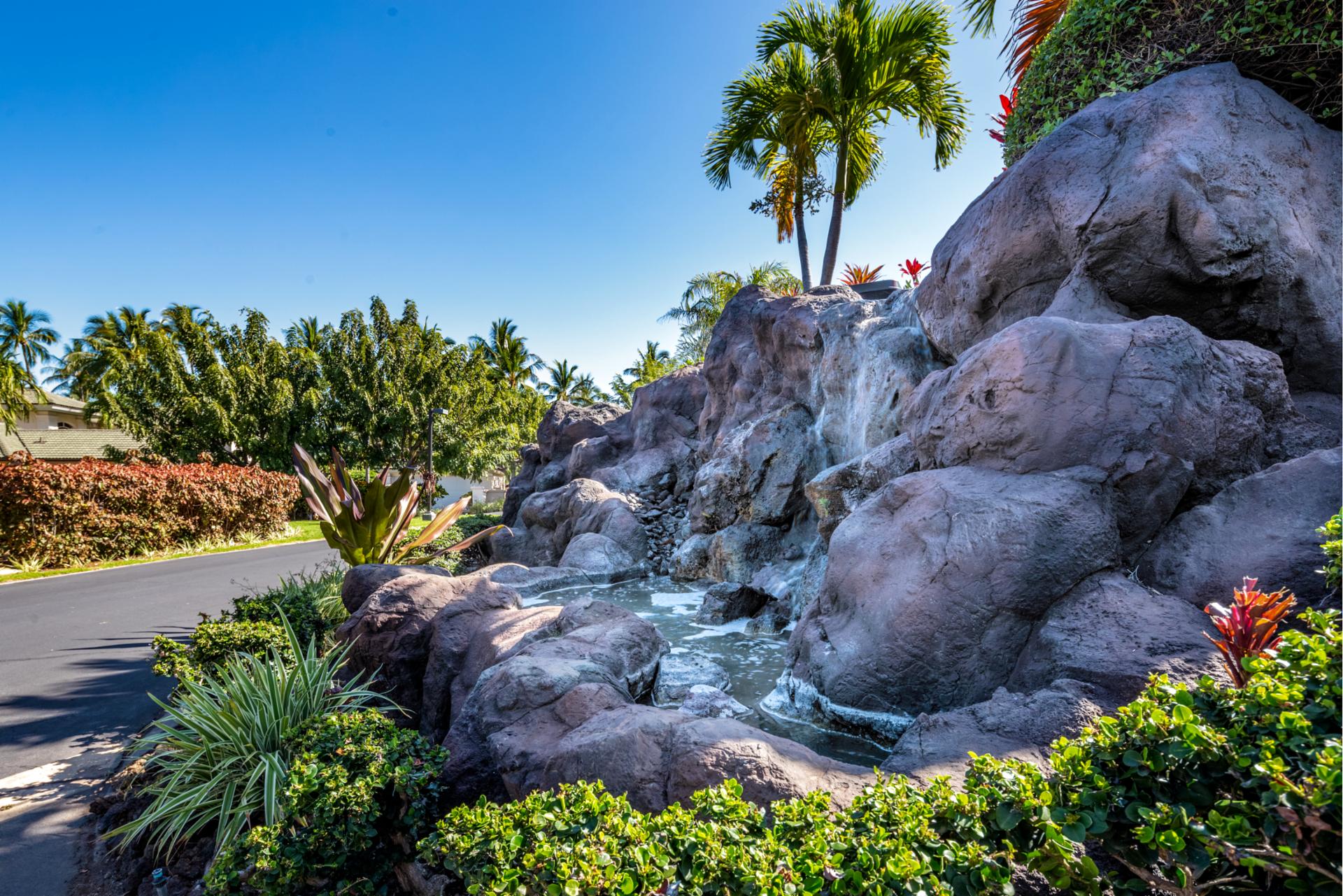 A tranquil landscaped garden featuring a cascading waterfall over large volcanic rocks, surrounded by lush greenery and tropical plants under a clear blue sky.