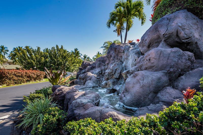 A tranquil landscaped garden featuring a cascading waterfall over large volcanic rocks, surrounded by lush greenery and tropical plants under a clear blue sky.