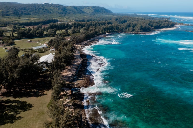 Aerial look at the rugged shoreline near Kuilima Estates, where turquoise waves crash against lava rock and forested cliffs frame Oahu’s wild North Shore.