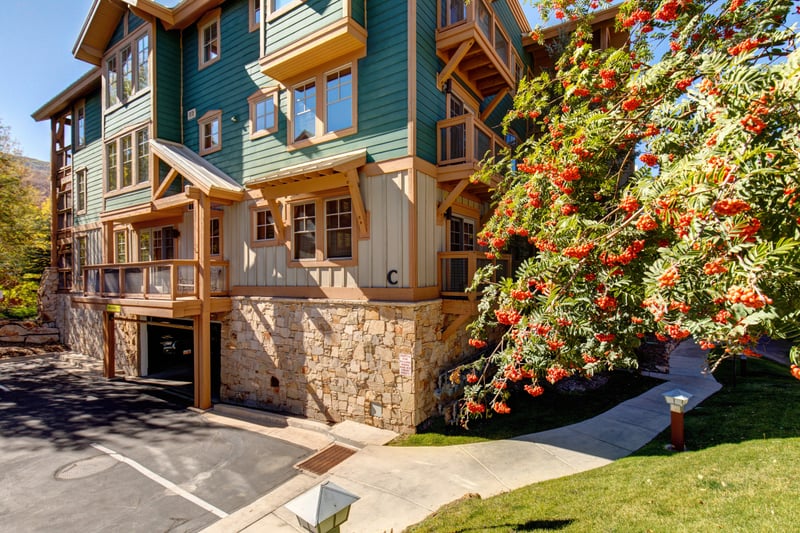 Charming mountain condo with lush landscaping, stone accents, and colorful autumn trees lining the drive.