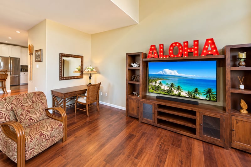 Living area with a wall unit holding a large TV and sound bar, plus a writing desk and patterned lounge chair.