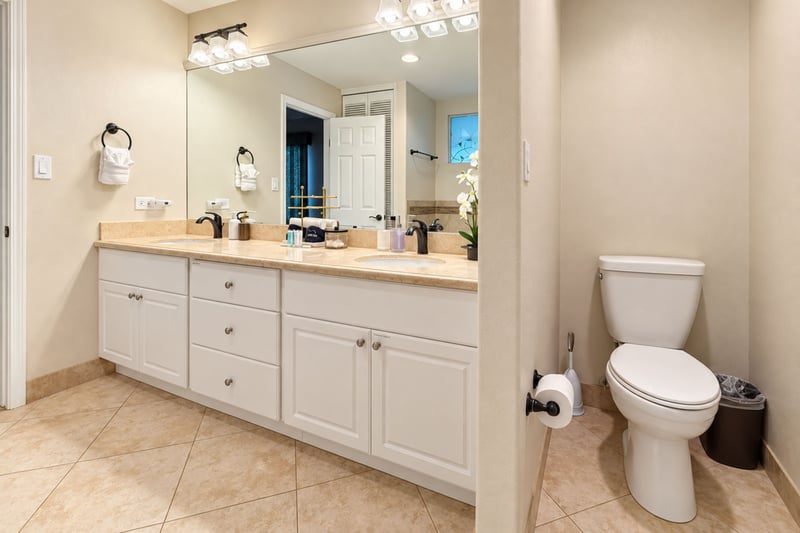 Primary bathroom vanity with two sinks and a separate toilet alcove, finished in warm cream tile and white cabinetry.