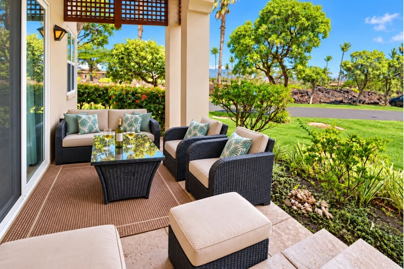 Covered ground-floor lanai with a cushioned wicker sofa, armchairs, and a glass top table, steps from green lawns and lush landscaping.