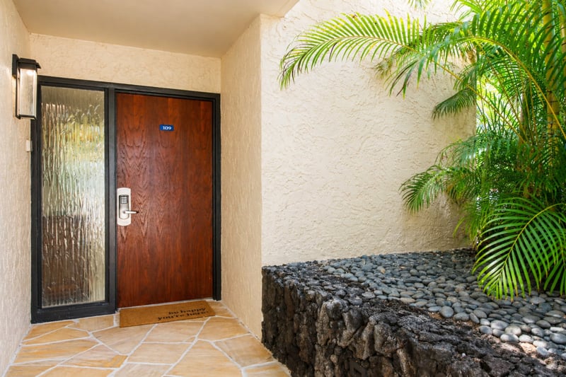 Keypad entry at Polo Beach Club 109, tucked beside lush palms and a lava-rock garden bed.