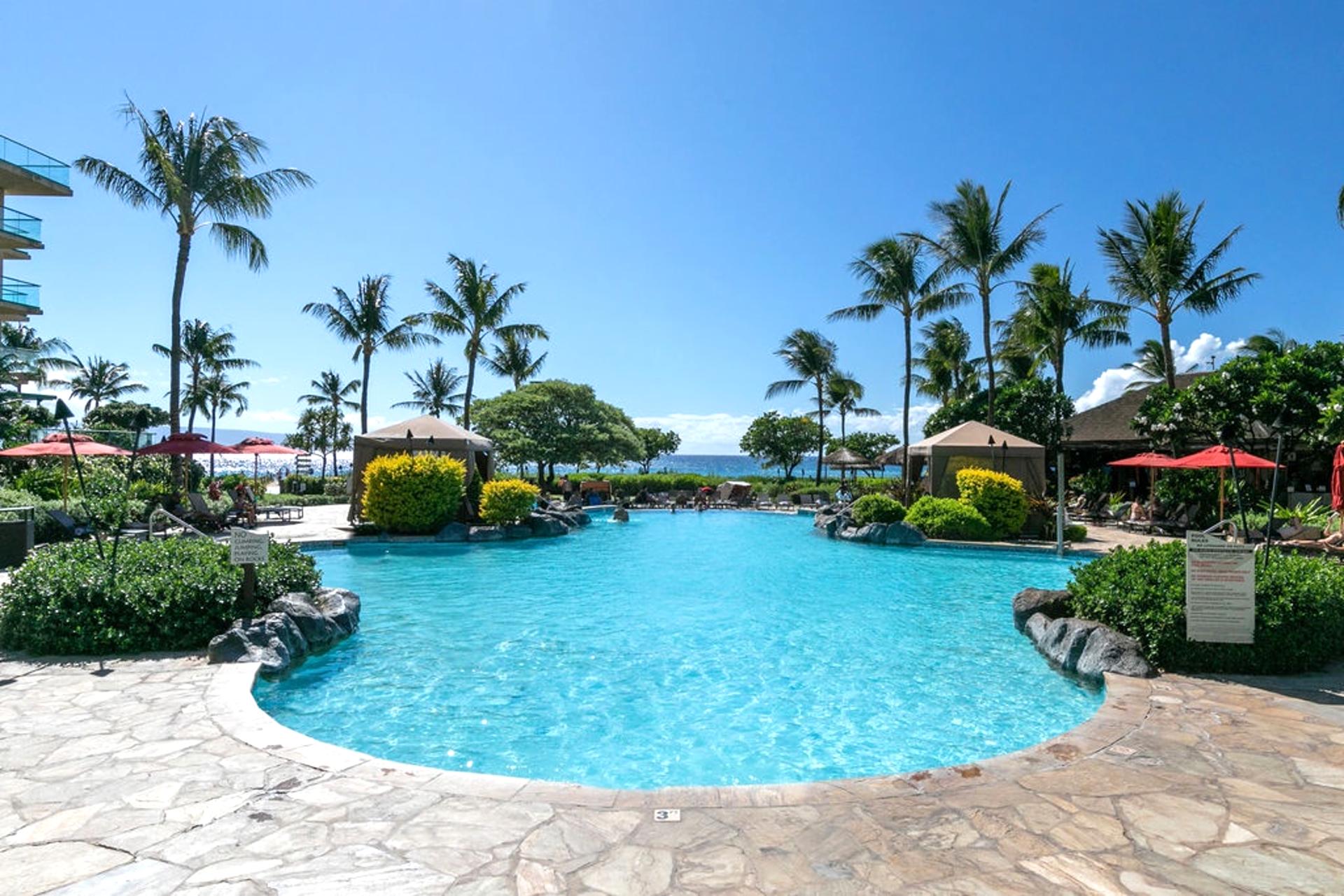 Resort-style pool at Honua Kai with lush landscaping, shaded cabanas, and a sparkling ocean view just beyond. Ideal for a refreshing dip or a full day in the sun.
