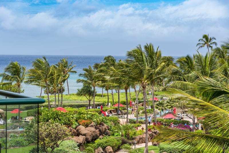 Tropical paradise unfolds below with palm-lined lawns, ocean views, and colorful cabanas along Maui’s scenic coastline