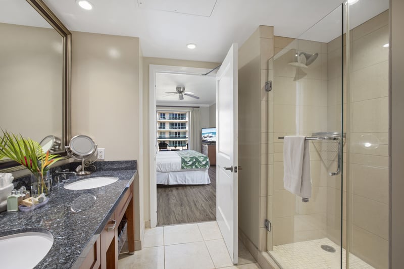Modern bathroom with glass shower, double sinks, and bedroom view.