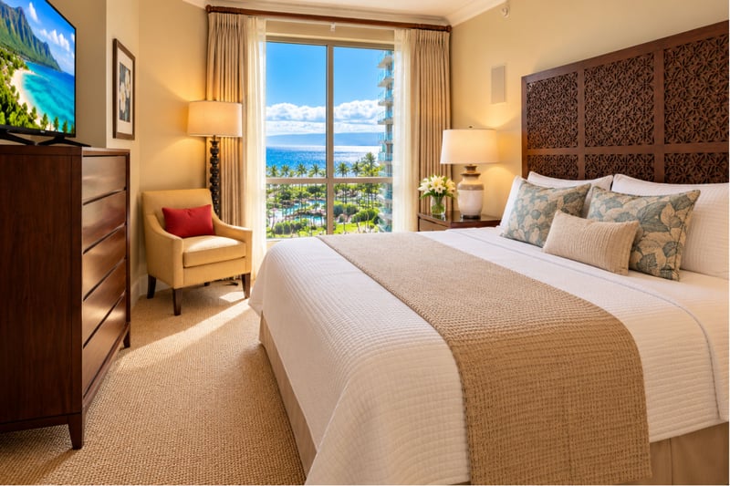 Pool and ocean views through the window frame this bedroom, with a king bed, a carved wood headboard, a chair and a television.
