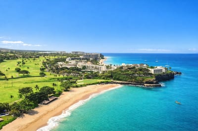 A breathtaking aerial view of Ka’anapali Beach glows under the setting sun, where lush fairways meet tranquil turquoise waters and luxury resorts line the coast in perfect harmony.