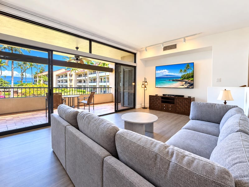 Your living room opens through wide sliding doors to a covered lanai with a table and chairs, palm trees, and a glimpse of the ocean, with a large TV above a wood console.