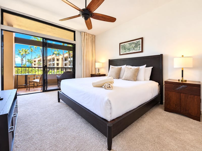 Your primary bedroom centers on a king bed with matching nightstands, with sliding doors opening to a lanai with palm views and a glimpse of the ocean.