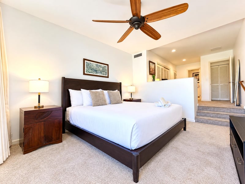 A few carpeted steps lead up from your primary bedroom, where a ceiling fan and framed island artwork sit above the king bed.