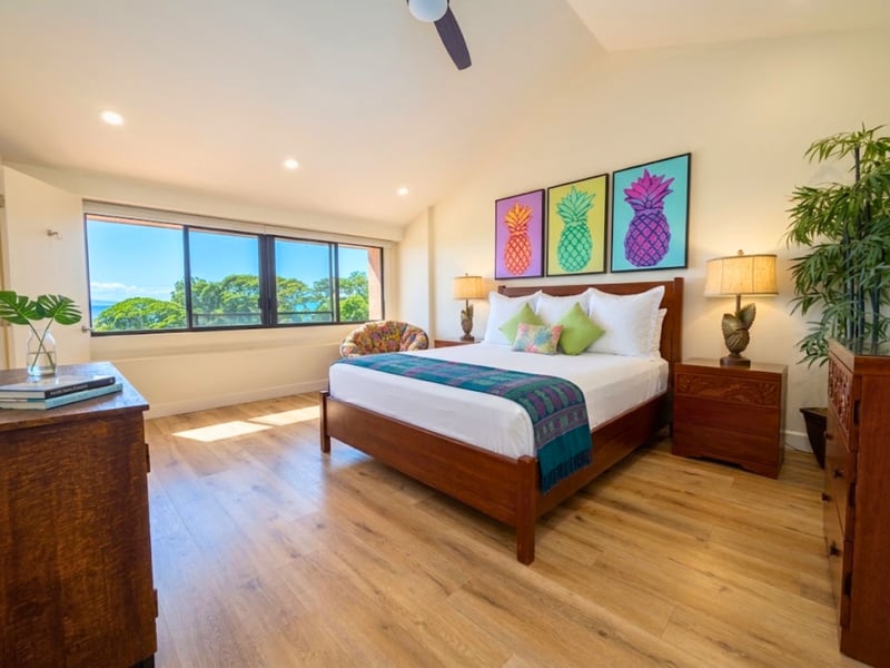 A trio of pineapple prints brightens the primary bedroom, where a vaulted ceiling and a wide window frame tree lined ocean views.