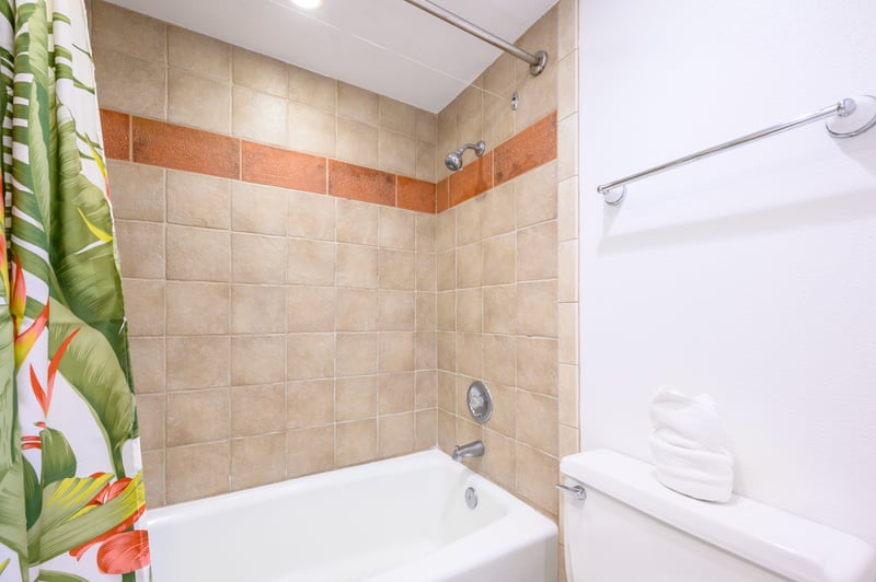 The tub and shower are framed in sandy-toned tile with a bold terracotta band, evoking natural textures and island tones