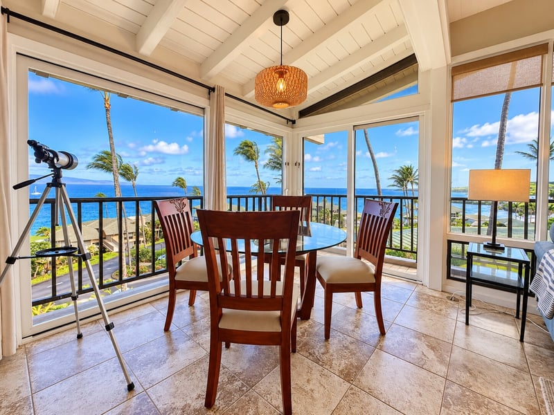 A round glass dining table seats four beneath a woven pendant light, with corner windows opening to ocean and palm views. KBV-15B3