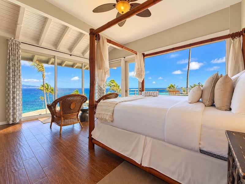 Wake to ocean views from a four-poster king bed, with a wicker seating pair and balcony access beside the corner windows. KBV-15B3