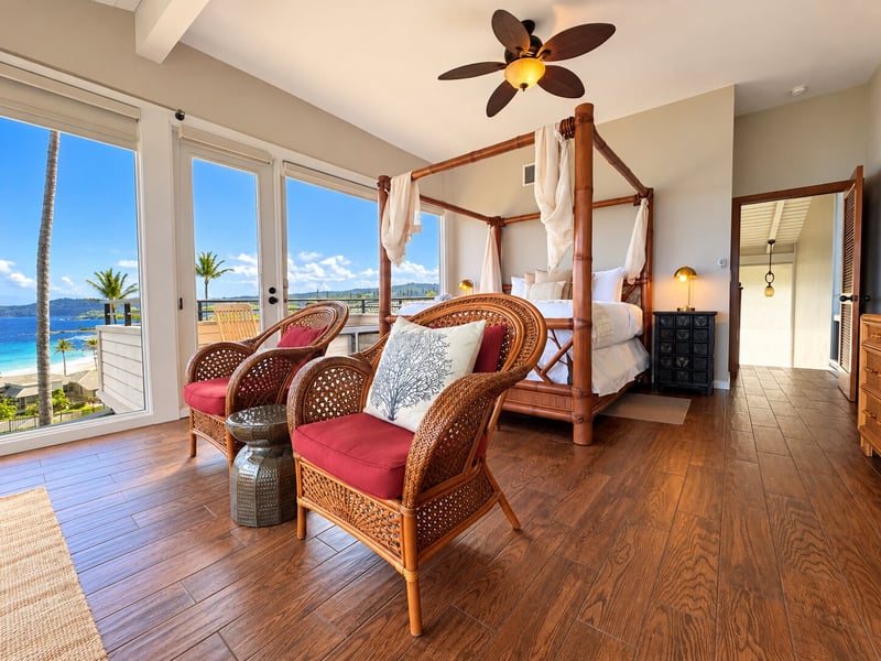 Wicker chairs and a side table sit near the glass doors, with the balcony and ocean beyond the four-poster bed. KBV-15B3