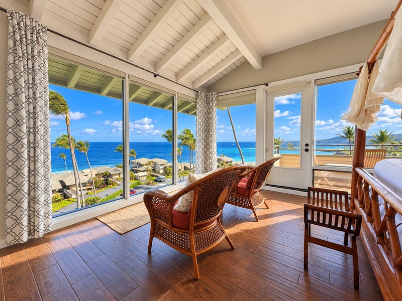 Corner windows wrap the bedroom sitting area, where two wicker chairs face the ocean beneath a beamed, sloped ceiling. KBV-15B3