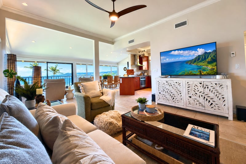 Your living room's wall-mounted TV sits opposite an open kitchen and dining area, with ocean views visible through the far windows.