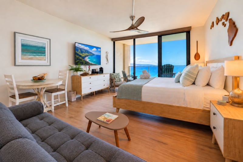 Open studio layout with a bed, tufted sofa, and dining table for two beneath a wall-mounted TV, all facing floor-to-ceiling ocean views MAH-1002