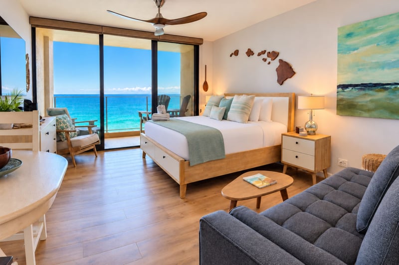 Sleeping and sitting area with Hawaiian island wall art, a ceiling fan, and sliding glass doors framing the blue Pacific MAH-1002