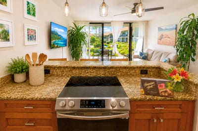 The glass-top range and open pass-through counter connect the kitchen straight to the living room and lanai beyond, where palm trees and the pool sparkle just past the sliding doors.