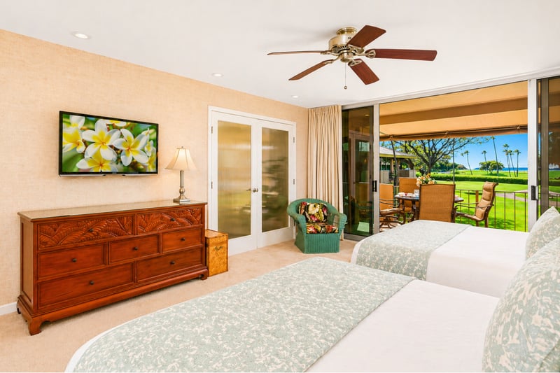 Guest bedroom dresser at Maui Eldorado H207 — wall-mounted TV, wicker armchair, and sliding-door access to the covered lanai