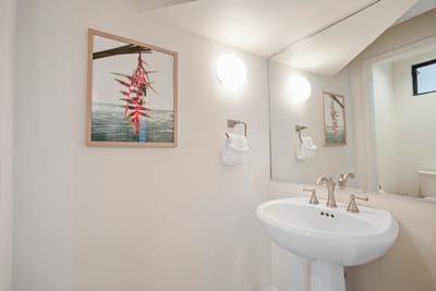 A bright and minimal guest bath featuring a pedestal sink, elegant fixtures, and coastal-inspired tropical wall art