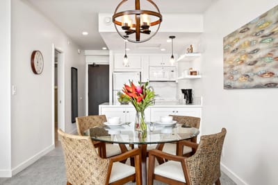 Gather around a glass-topped table framed by woven chairs, soft coastal tones, and warm globe pendant lighting