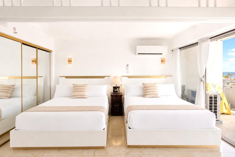 Two crisp queen beds dressed in white linen with warm gold accents, split A/C for cool comfort, and sliding doors opening to the lanai and a peek of blue Pacific.