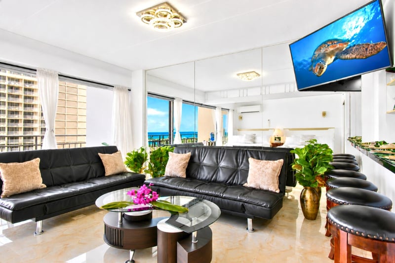 Comfortable dual-sofa living area beneath a modern chandelier, Smart TV, and a straight-ahead ocean view - the welcoming heart of this open Ilikai suite.
