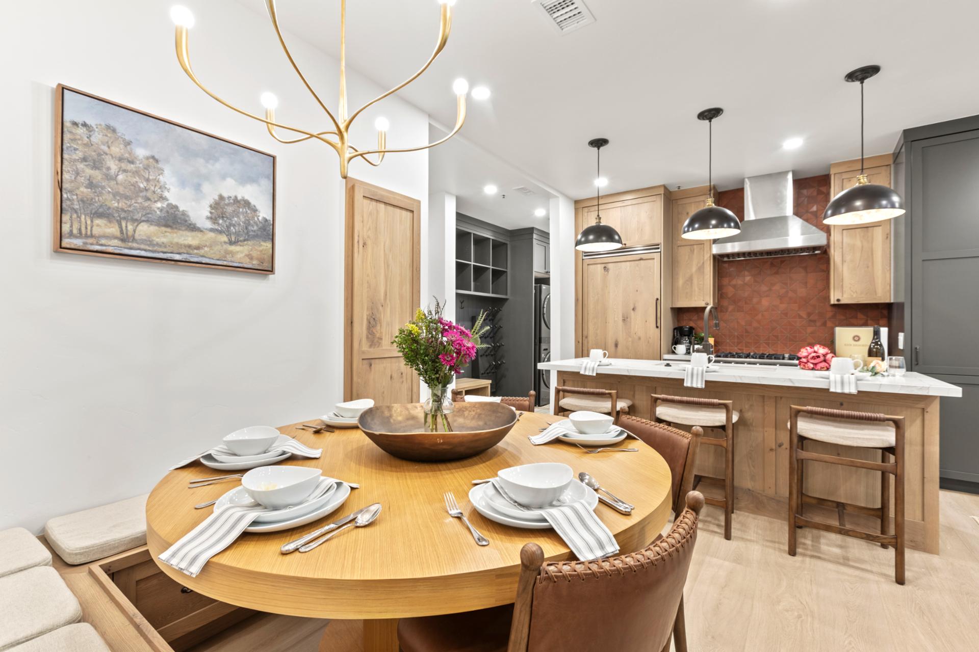 A round dining table for four sits beneath a sculptural light fixture, with views into the modern kitchen.