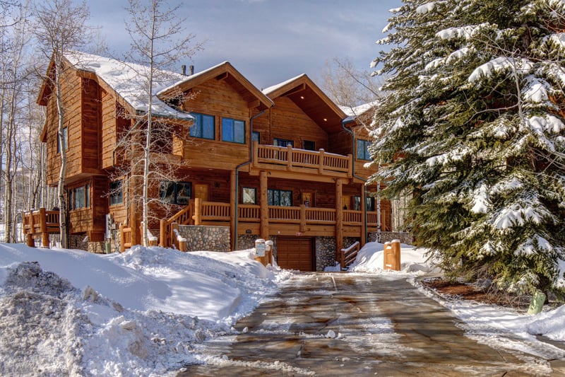 Charming log cabin with snowy driveway, nestled among winter pines. TWL-5B