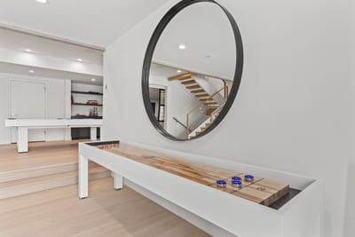 Modern entertainment space with white shuffleboard, wood accents, circular mirror.