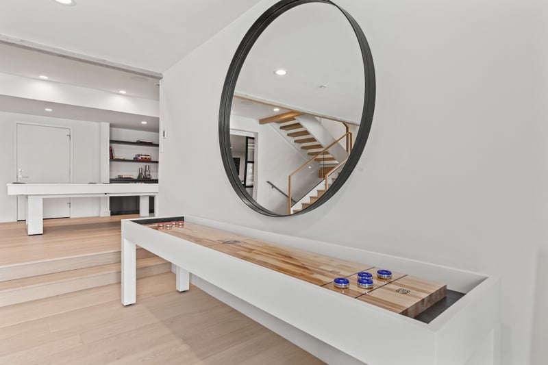 Modern entertainment space with white shuffleboard, wood accents, circular mirror.