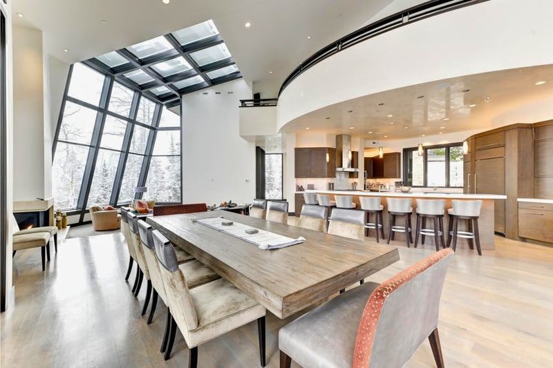 Great room dining table for ten beneath a dramatic angled glass wall, open to the kitchen island and a snowy forest view.