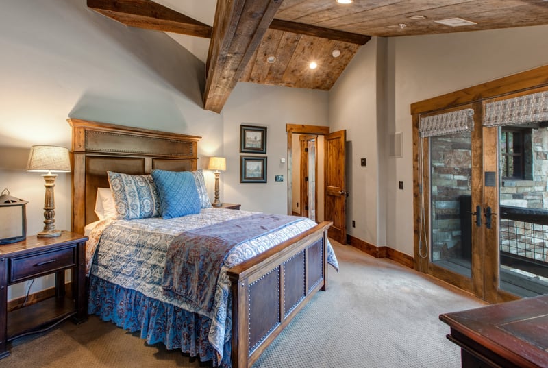 Guest Bedroom 1 features a King bed, exposed beams, balcony doors, and cozy lighting for a restful mountain stay.