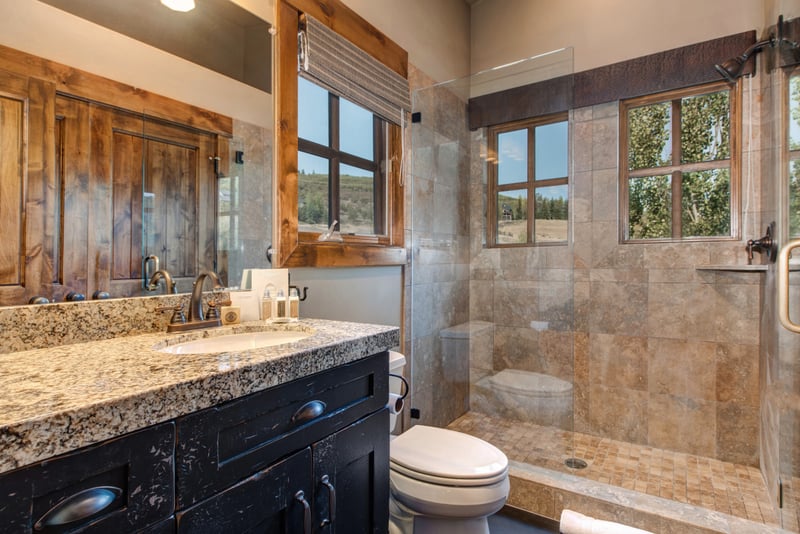 Enjoy a private ensuite with a granite vanity, glass walk-in shower, and natural light through rustic wood-trimmed windows.